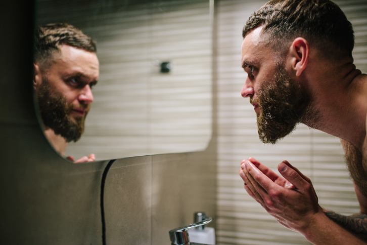 A man with a full beard looks into a bathroom mirror 
